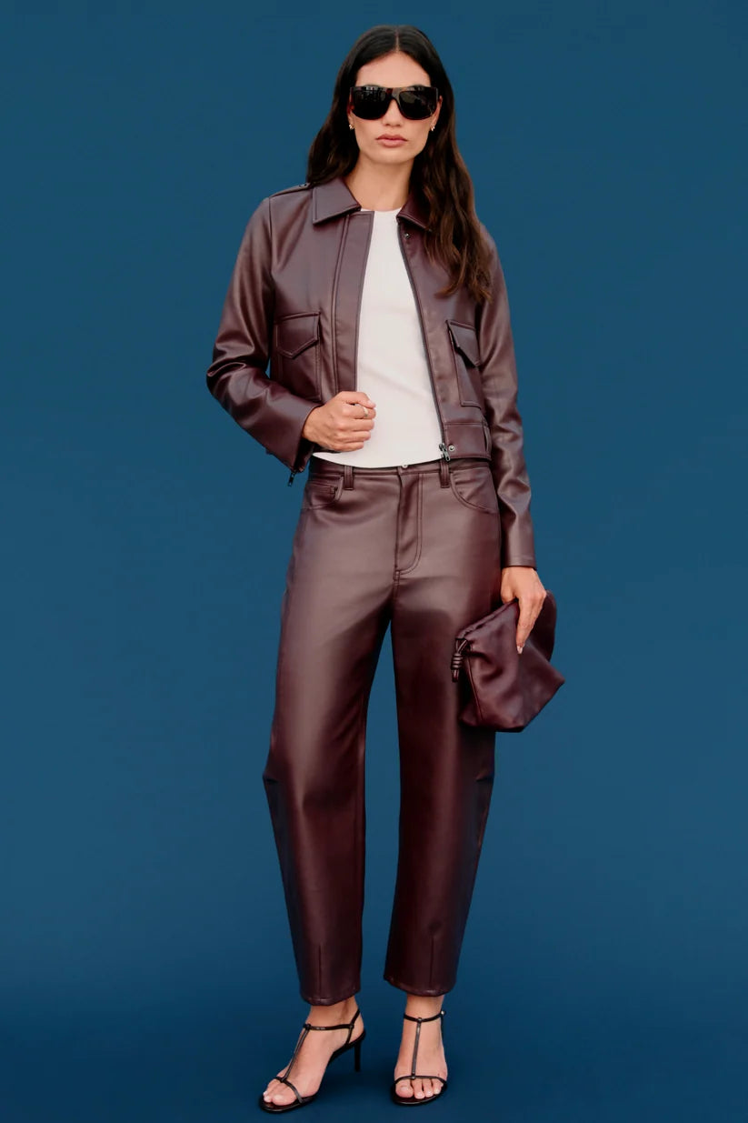 A model stands against a solid blue studio background wearing a matching brown faux-leather jacket and trousers set, styled with a white t-shirt, black sunglasses, and black strappy heels.