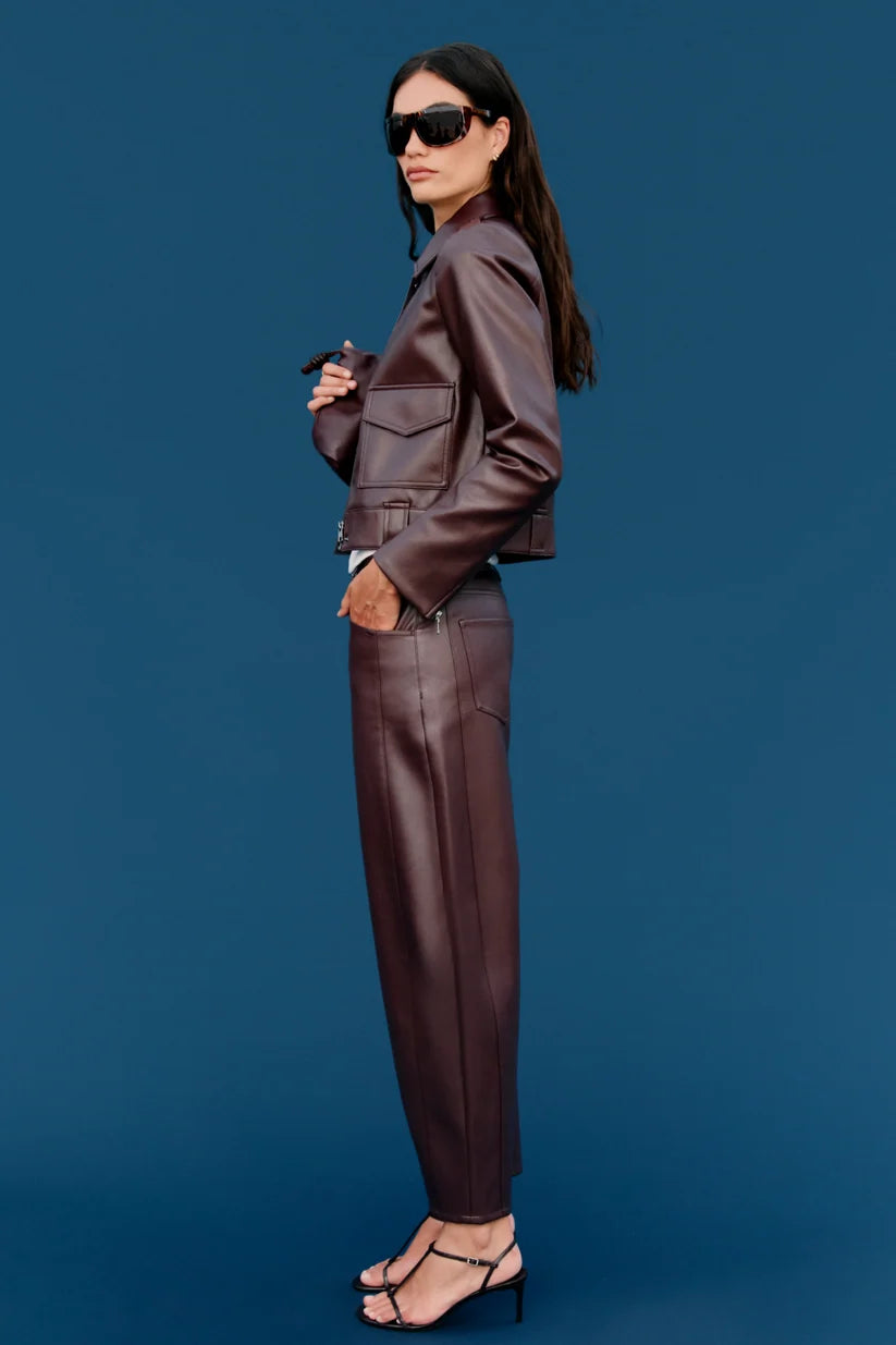 A model wearing a matching dark brown faux leather jacket and trousers set, styled with black sunglasses and black strappy heels, standing against a solid blue background.