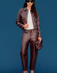 A model stands against a solid blue studio background wearing a matching brown faux-leather jacket and trousers set, styled with a white t-shirt, black sunglasses, and black strappy heels.