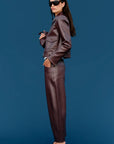 A model wearing a matching dark brown faux leather jacket and trousers set, styled with black sunglasses and black strappy heels, standing against a solid blue background.