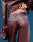 A close-up back view of a model wearing dark brown faux leather pants and a matching cropped jacket, holding a matching brown clutch bag against a solid blue background.