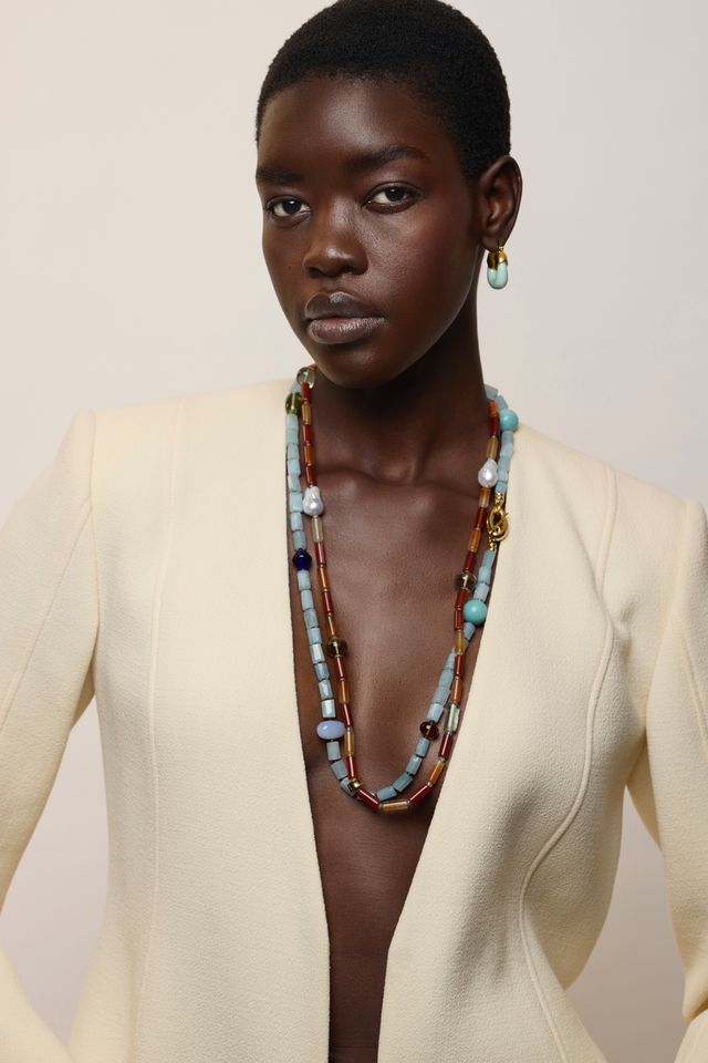 A model wearing a cream-colored blazer, Mini Organic Hoops in Seafoam and a long, multi-strand beaded necklace with turquoise, blue, brown, and gold beads, set against a plain beige background.