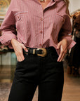 A person wearing a pink and white vertically striped button-up shirt tucked into black jeans with a black belt featuring a large gold buckle, standing in a softly blurred indoor setting.