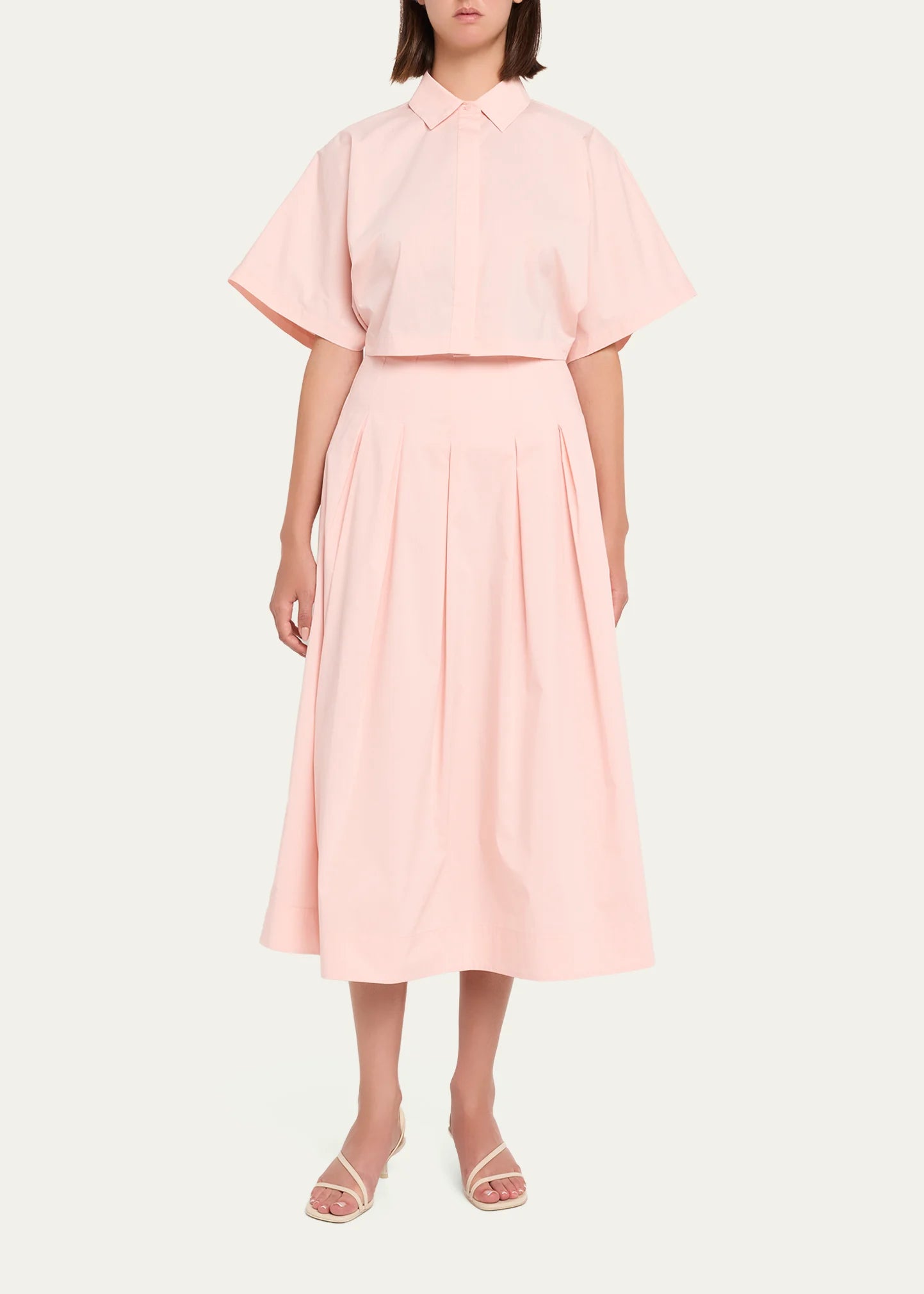 A model wearing a light pink two-piece set consisting of a short-sleeve collared blouse and a matching pleated midi skirt, paired with white heeled sandals, against a solid off-white background.