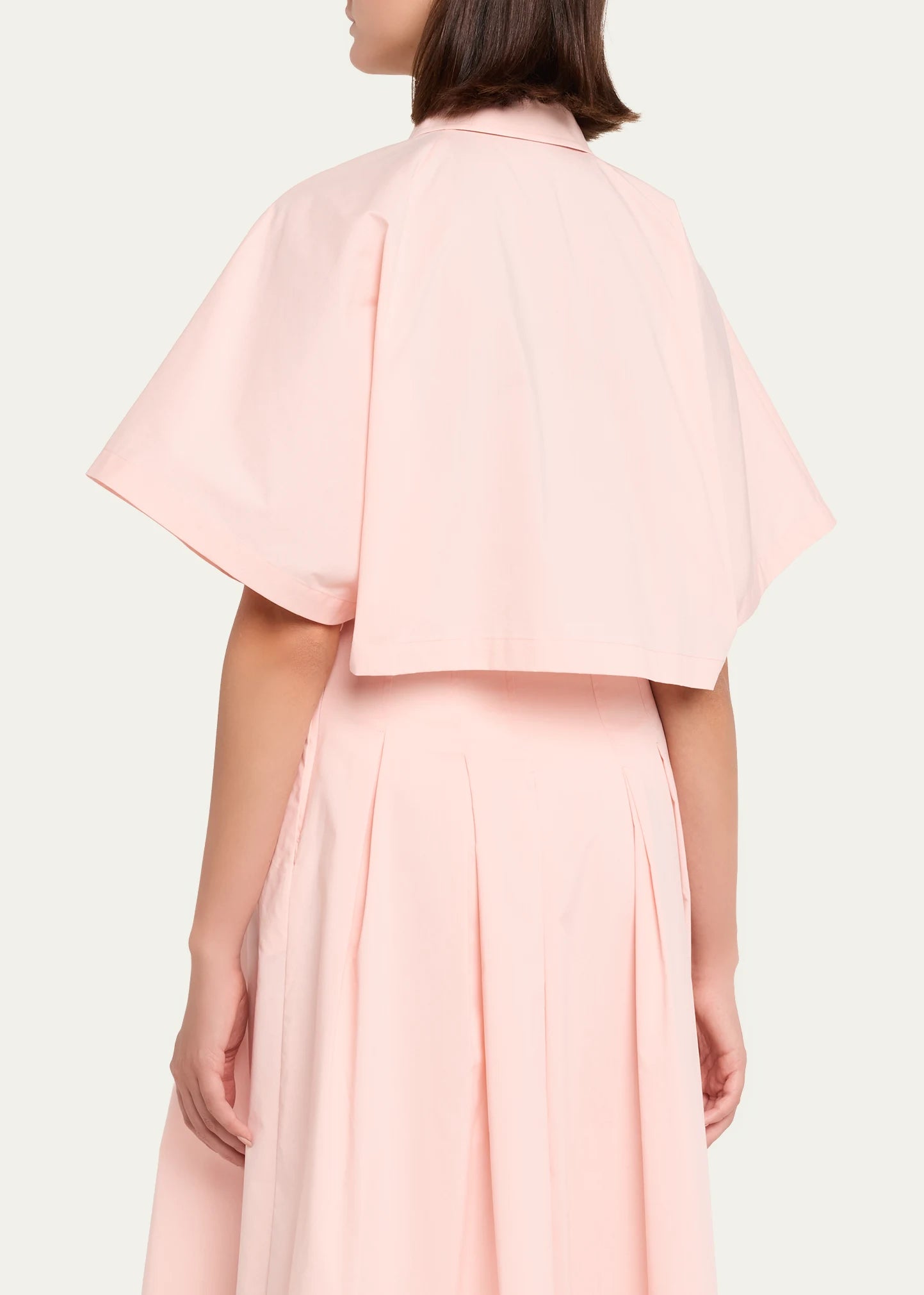 A model shown from the back wearing a light pink two-piece outfit consisting of a short-sleeved cropped top with a collar and a matching pleated skirt, against a plain off-white background.