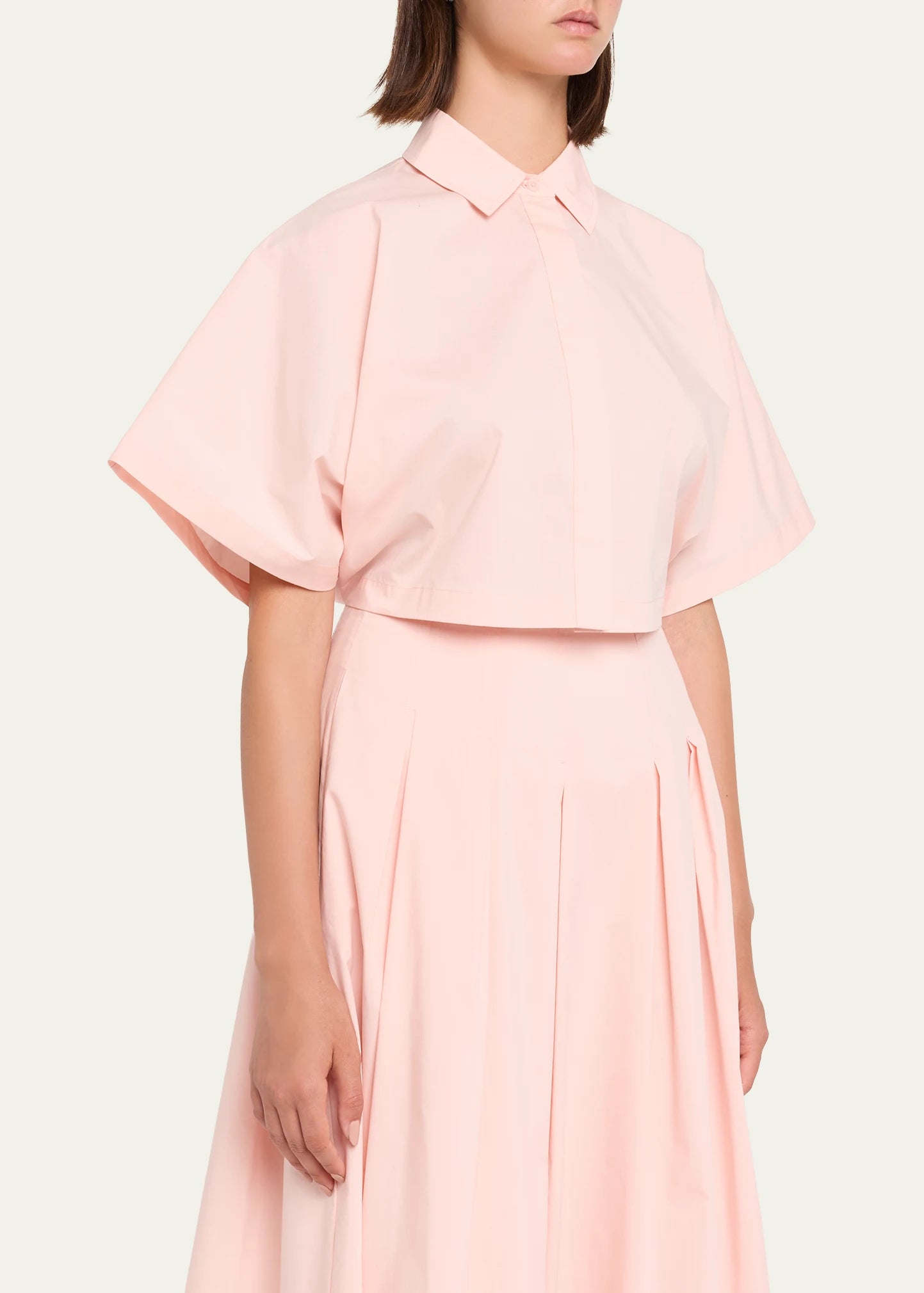 A model wearing a light pink two-piece set consisting of a short-sleeve collared blouse and a matching pleated skirt, shot against a solid off-white background.