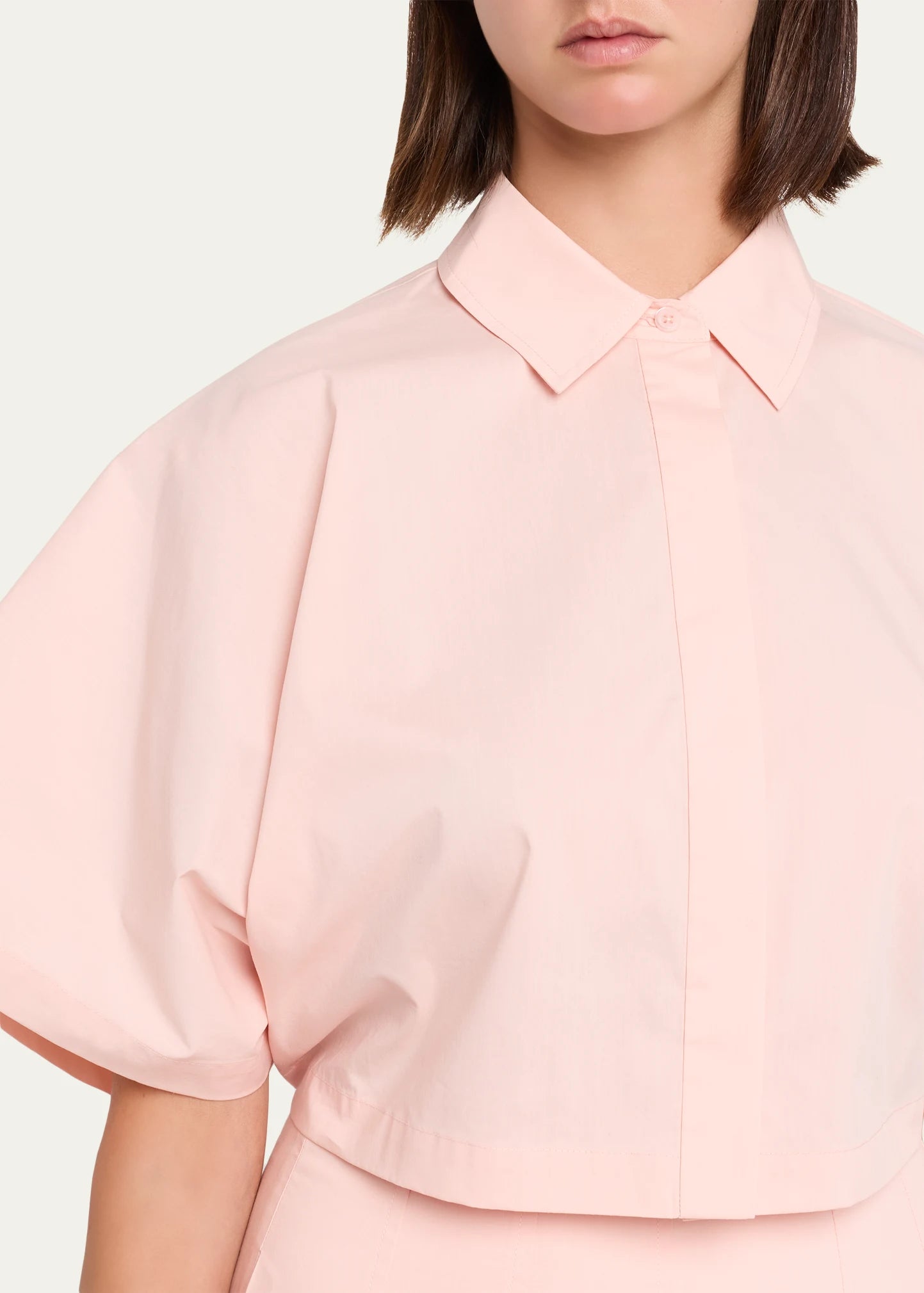 A close-up of a model wearing a light pink cropped blouse with voluminous short puff sleeves, a pointed collar, and a button front, set against a plain off-white background.