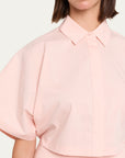 A close-up of a model wearing a light pink cropped blouse with voluminous short puff sleeves, a pointed collar, and a button front, set against a plain off-white background.
