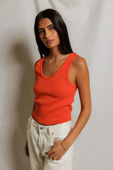 Woman wearing an orange tank top and white pants against a plain background