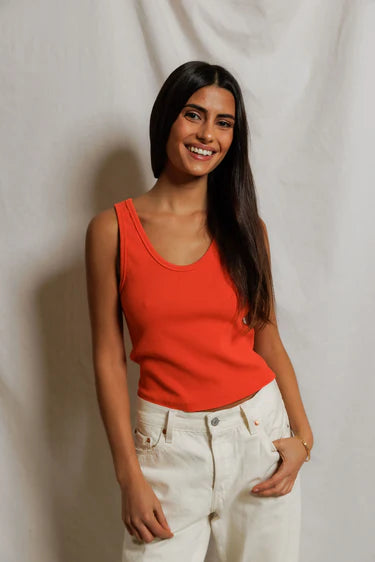 Woman wearing an orange tank top and white pants against a plain background
