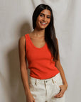 Woman wearing an orange tank top and white pants against a plain background