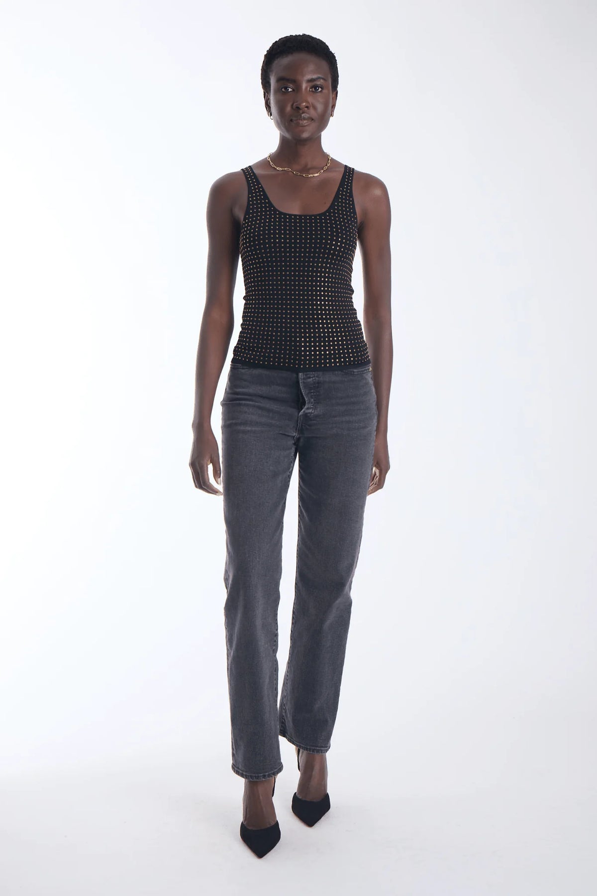 A model stands against a white studio background wearing a black sleeveless top with a small metallic stud pattern, dark gray jeans, and black pointed heels.