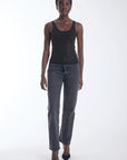 A model stands against a white studio background wearing a black sleeveless top with a small metallic stud pattern, dark gray jeans, and black pointed heels.