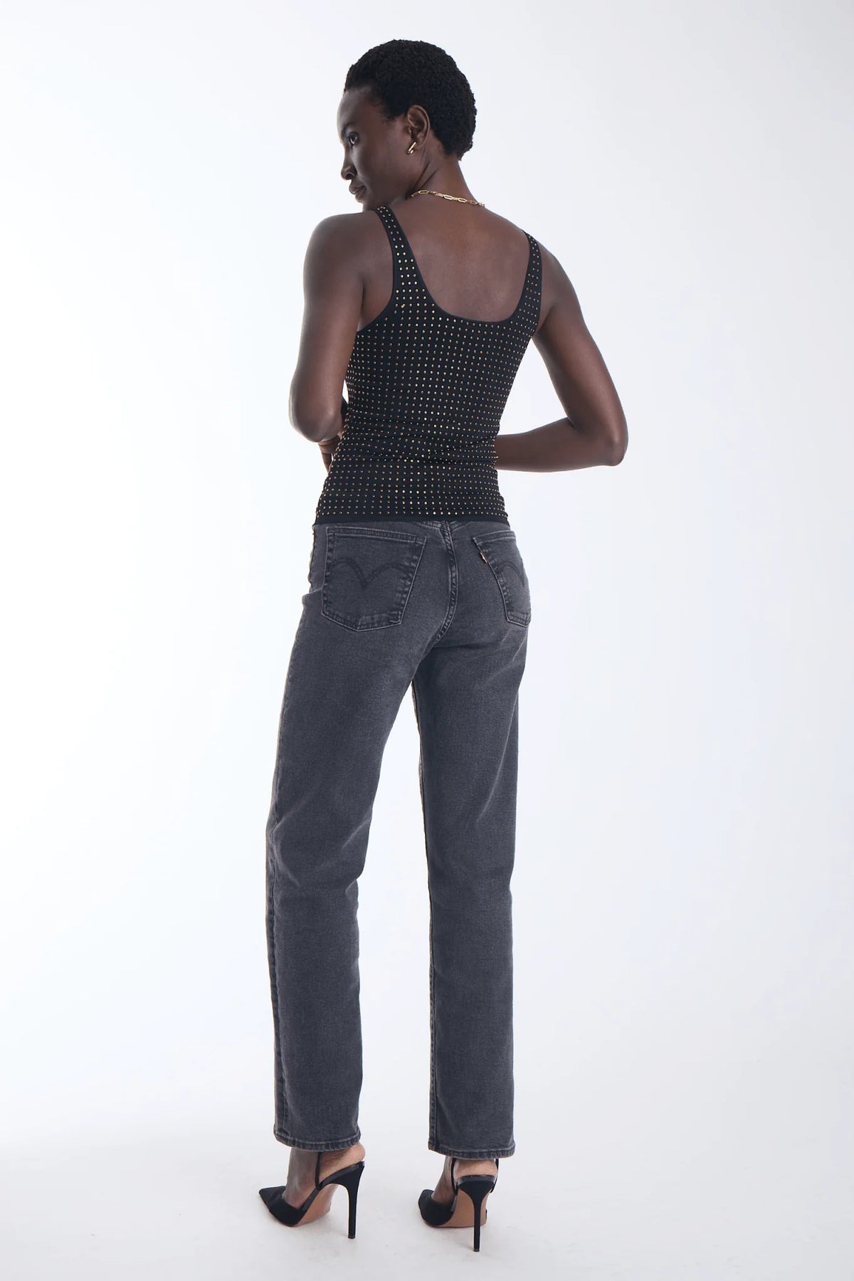 A model is shown from the back wearing a black studded halter top, dark gray jeans, and black high heels against a plain white studio background.