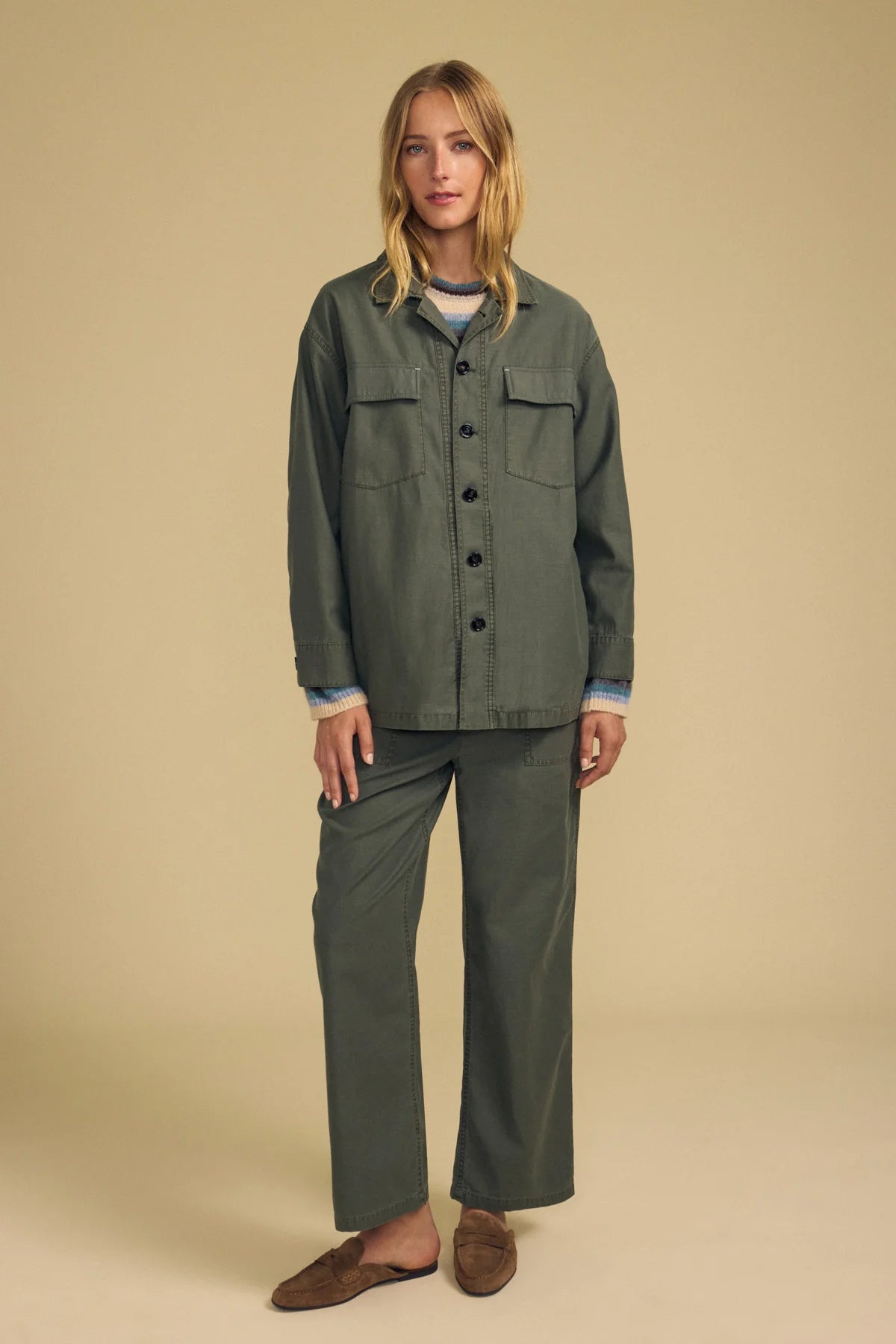 A woman modeling a matching two-piece olive green set consisting of a long-sleeve button-front shirt jacket and wide-leg trousers, styled with a light blue striped shirt and brown slip-on shoes against a solid beige studio background.