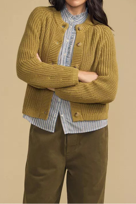 A woman modeling a mustard yellow ribbed knit cardigan over a blue and white striped shirt, paired with olive green pants, against a solid beige background.