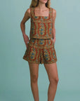 A woman modeling a matching sleeveless top and shorts set with a rust-orange, blue, and beige paisley pattern, paired with black slip-on shoes, against a solid light blue background.