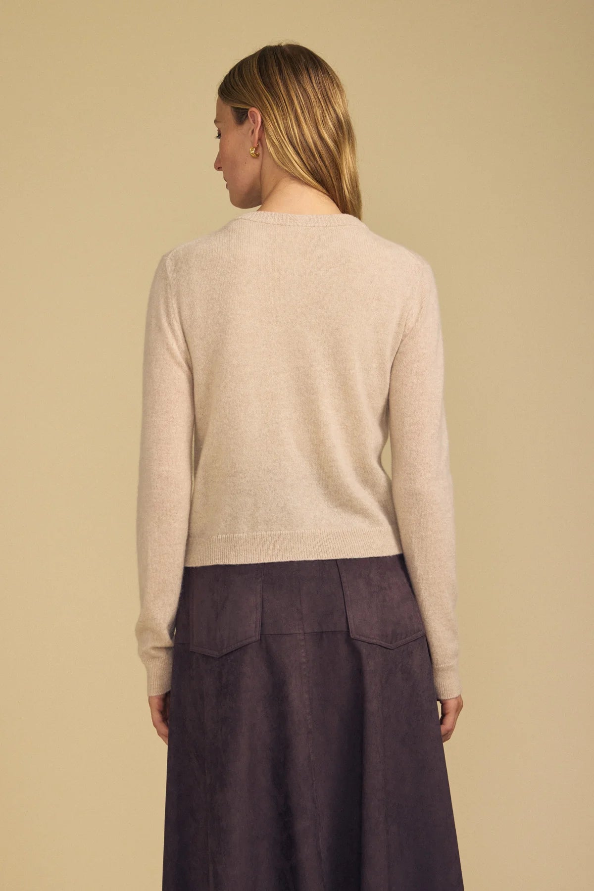 A woman shown from the back wearing a light beige knit sweater and a dark purple skirt, set against a solid warm beige background.