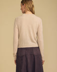 A woman shown from the back wearing a light beige knit sweater and a dark purple skirt, set against a solid warm beige background.