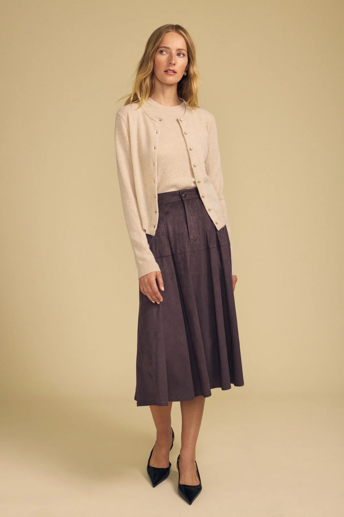 A woman modeling a light beige cardigan over a cream top and a dark purple pleated midi skirt, paired with black pointed-toe heels, against a solid beige studio background.