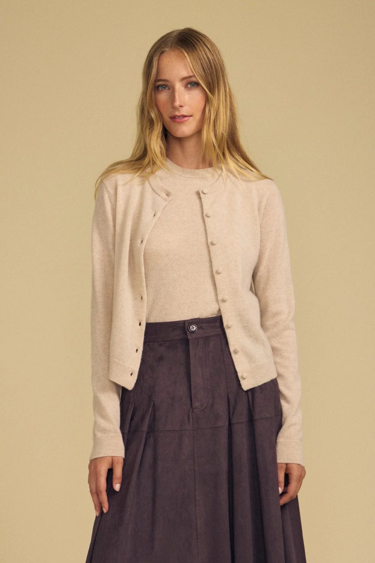 A woman modeling a light beige knit cardigan with decorative buttons over a matching beige top and a dark brown skirt, set against a solid beige background.