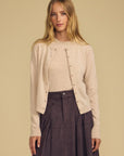 A woman modeling a light beige knit cardigan with decorative buttons over a matching beige top and a dark brown skirt, set against a solid beige background.