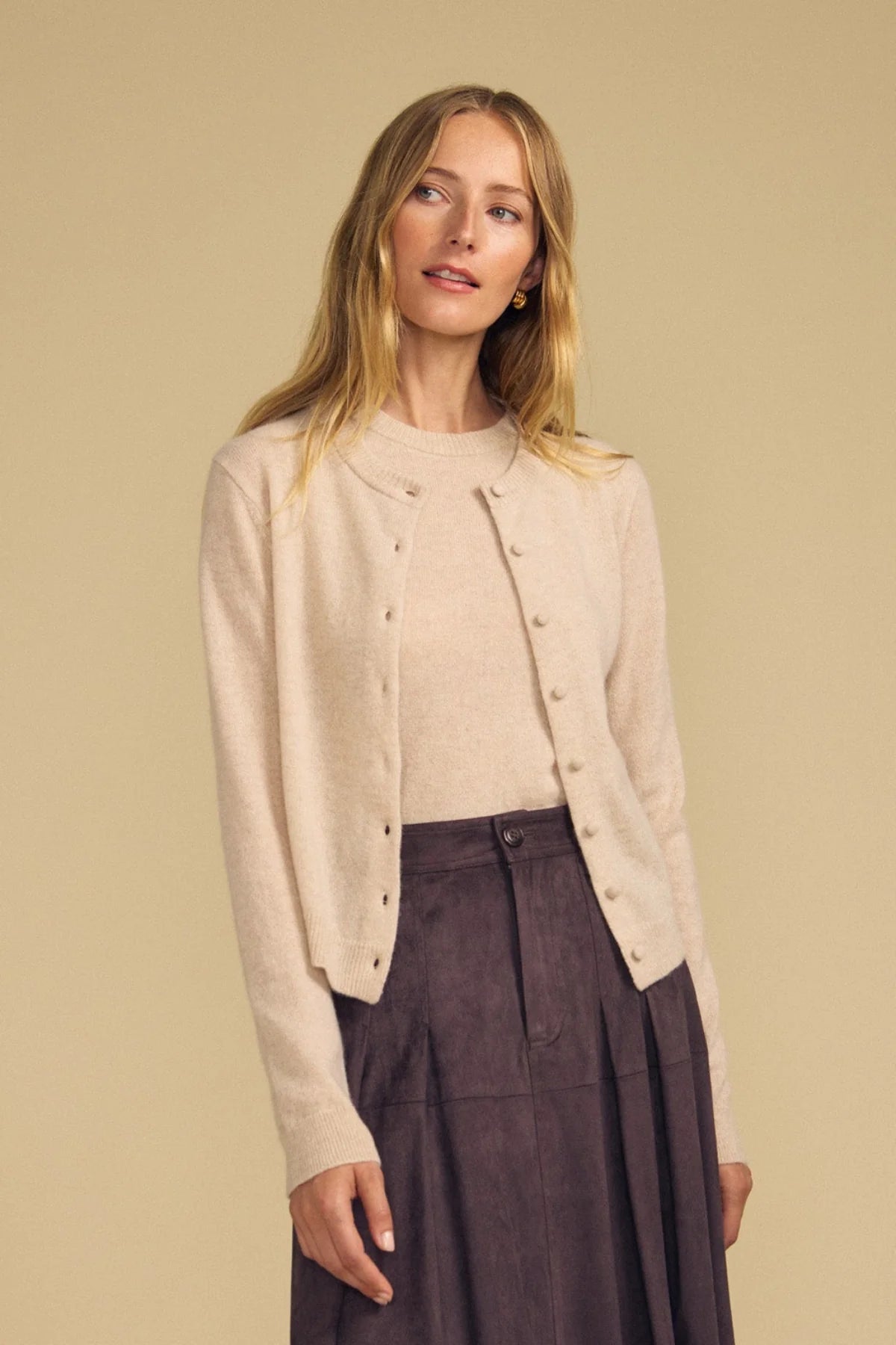 A woman modeling a light beige knit cardigan with decorative buttons over a matching beige top and a dark brown pleated skirt, set against a solid beige background.