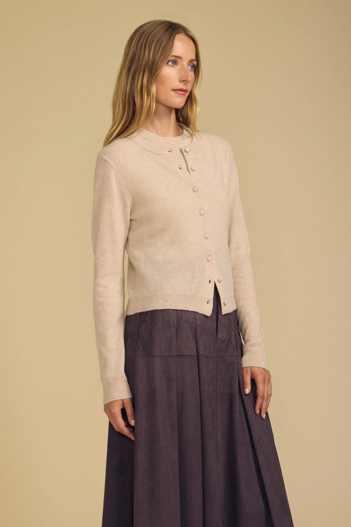 A woman modeling a light beige knit cardigan with a button front and a dark mauve pleated skirt against a solid beige background.