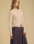 A woman modeling a light beige knit cardigan with a button front and a dark mauve pleated skirt against a solid beige background.