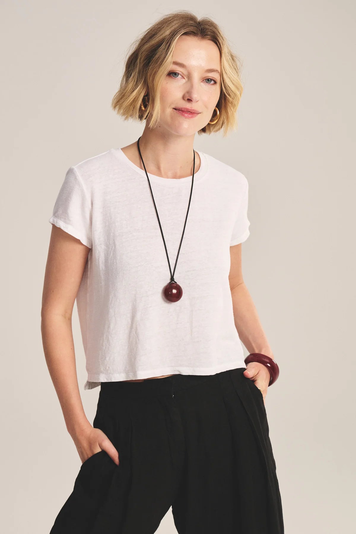 Woman wearing a white t-shirt and black pants with a long necklace against a plain background