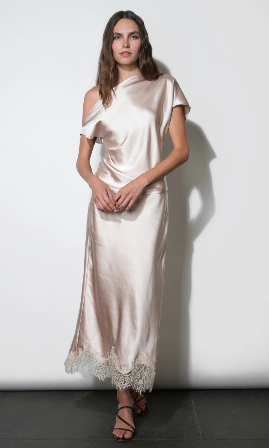 A woman models a champagne-colored satin midi dress with a draped, one-shoulder style and a lace-trimmed hem, set against a plain white wall.