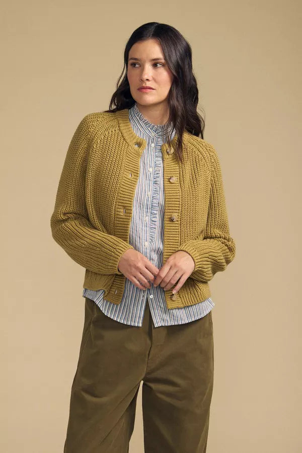 A woman modeling a mustard yellow ribbed knit cardigan with brown buttons, worn over a blue and white striped shirt and paired with olive green pants, against a solid beige background.