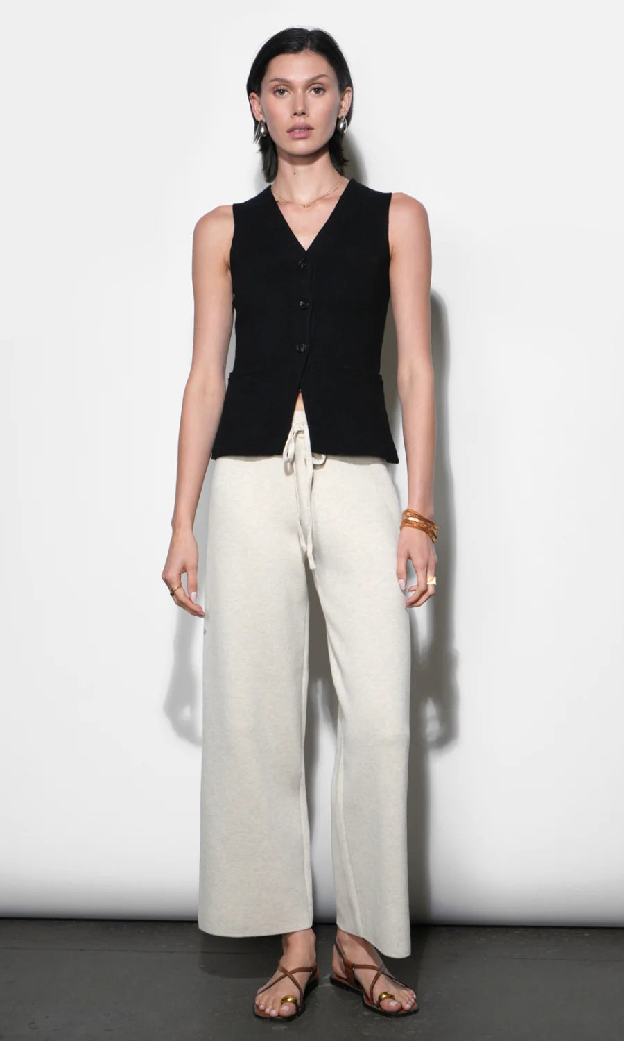 Woman wearing a black sleeveless vest and white pants against a white background