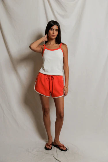 Woman wearing a white tank top with orange trim and orange shorts against a plain background