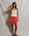 Woman wearing a white tank top with orange trim and orange shorts against a plain background