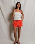 Woman wearing a white tank top with orange trim and orange shorts against a plain background