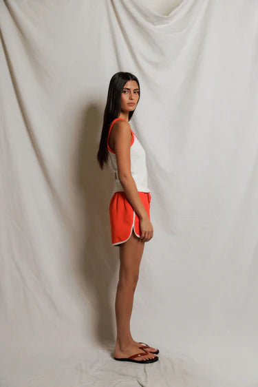 Woman wearing a white tank top with orange trim and orange shorts against a plain background, side view