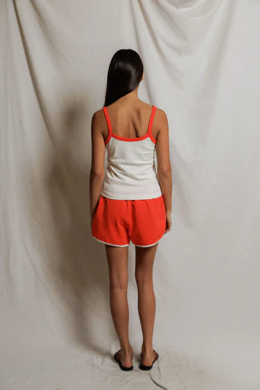 Woman wearing a white tank top with orange trim and orange shorts against a plain background, back view