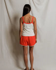Woman wearing a white tank top with orange trim and orange shorts against a plain background, back view