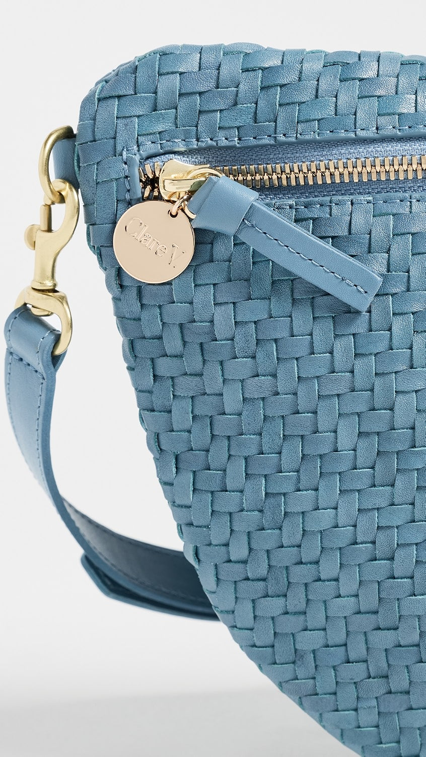 A close-up of a blue woven leather crossbody bag with a gold-tone zipper, a round metal brand tag, and a matching blue strap against a light gray background.