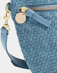 A close-up of a blue woven leather crossbody bag with a gold-tone zipper, a round metal brand tag, and a matching blue strap against a light gray background.