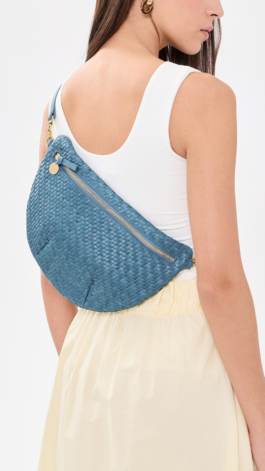 A woman wearing a white tank top and cream skirt carries a blue woven crossbody bag with gold hardware.