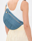 A woman wearing a white tank top and cream skirt carries a blue woven crossbody bag with gold hardware.