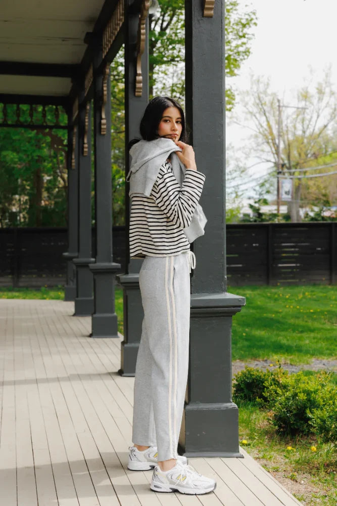Woman standing on a wooden porch wearing a striped sweater and light pants.