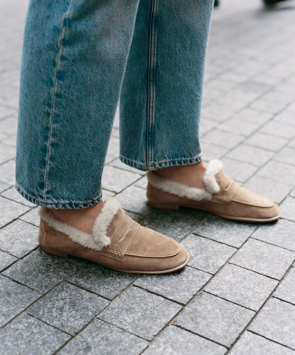 A person wearing beige suede slip-on loafers with white shearling lining, paired with blue jeans, standing on a gray paved outdoor surface.