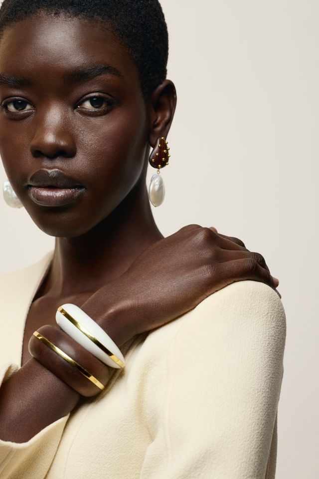 A close-up portrait of a model wearing a cream-colored wide cuff bracelet with gold trim on her wrist, along with matching cream and gold earrings, against a light beige background.