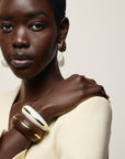 A close-up portrait of a model wearing a cream-colored wide cuff bracelet with gold trim on her wrist, along with matching cream and gold earrings, against a light beige background.