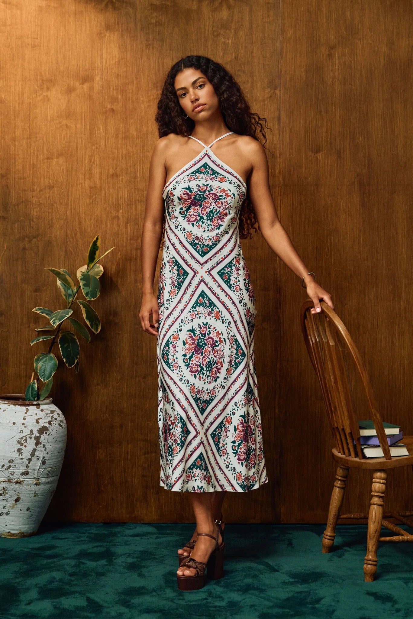 A woman models a sleeveless halter midi dress with a colorful floral and geometric diamond pattern, standing in front of a wooden wall next to a wooden chair and a potted plant.