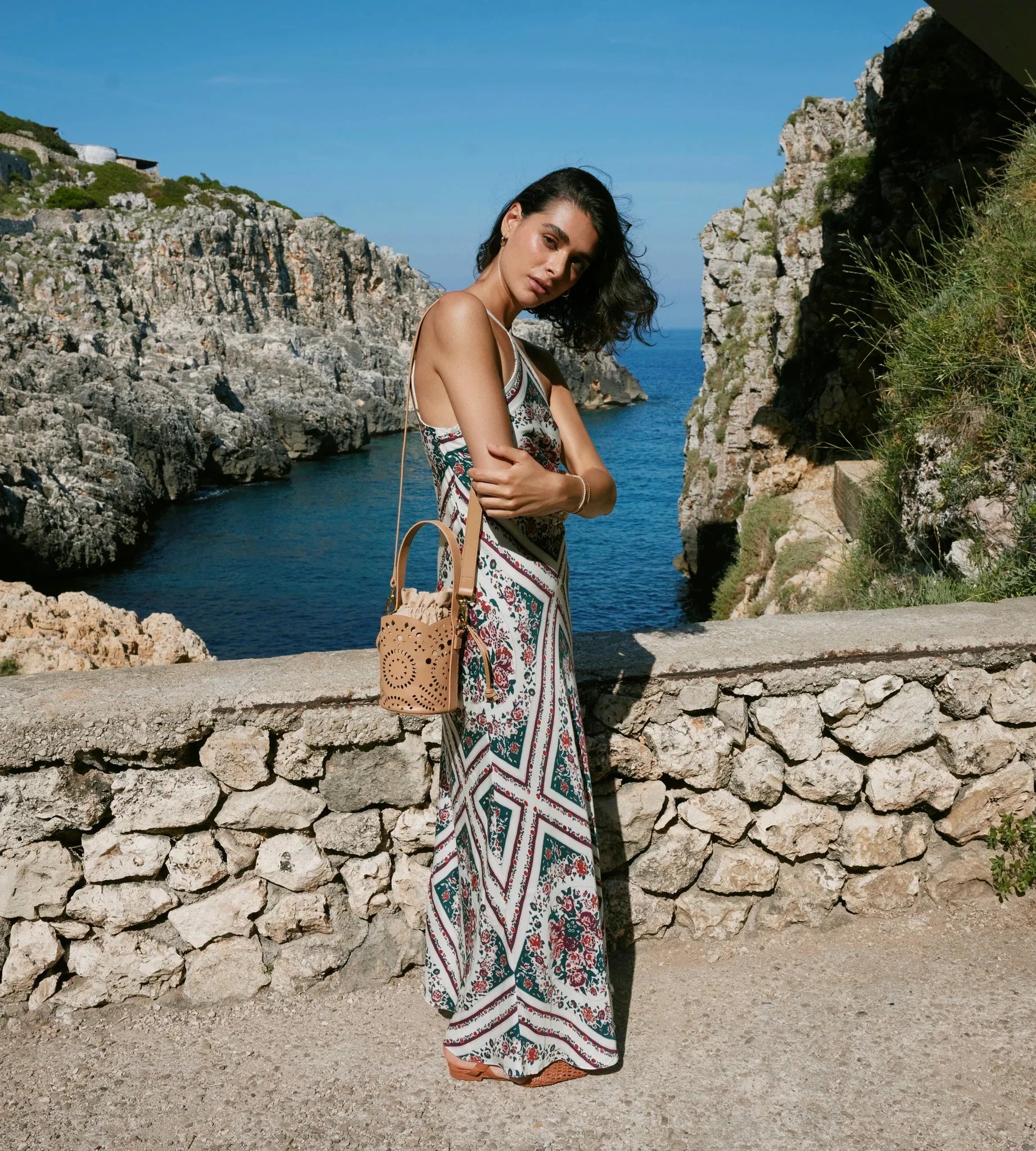 A woman stands outdoors by a rocky coastal cliffside overlooking the sea, wearing a sleeveless geometric print maxi dress and carrying a woven tan handbag.