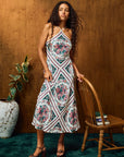A woman models a sleeveless midi dress with a white base and a colorful geometric floral pattern in pink, green, and blue, standing in a room with a wooden wall, a wooden chair, and a potted plant.